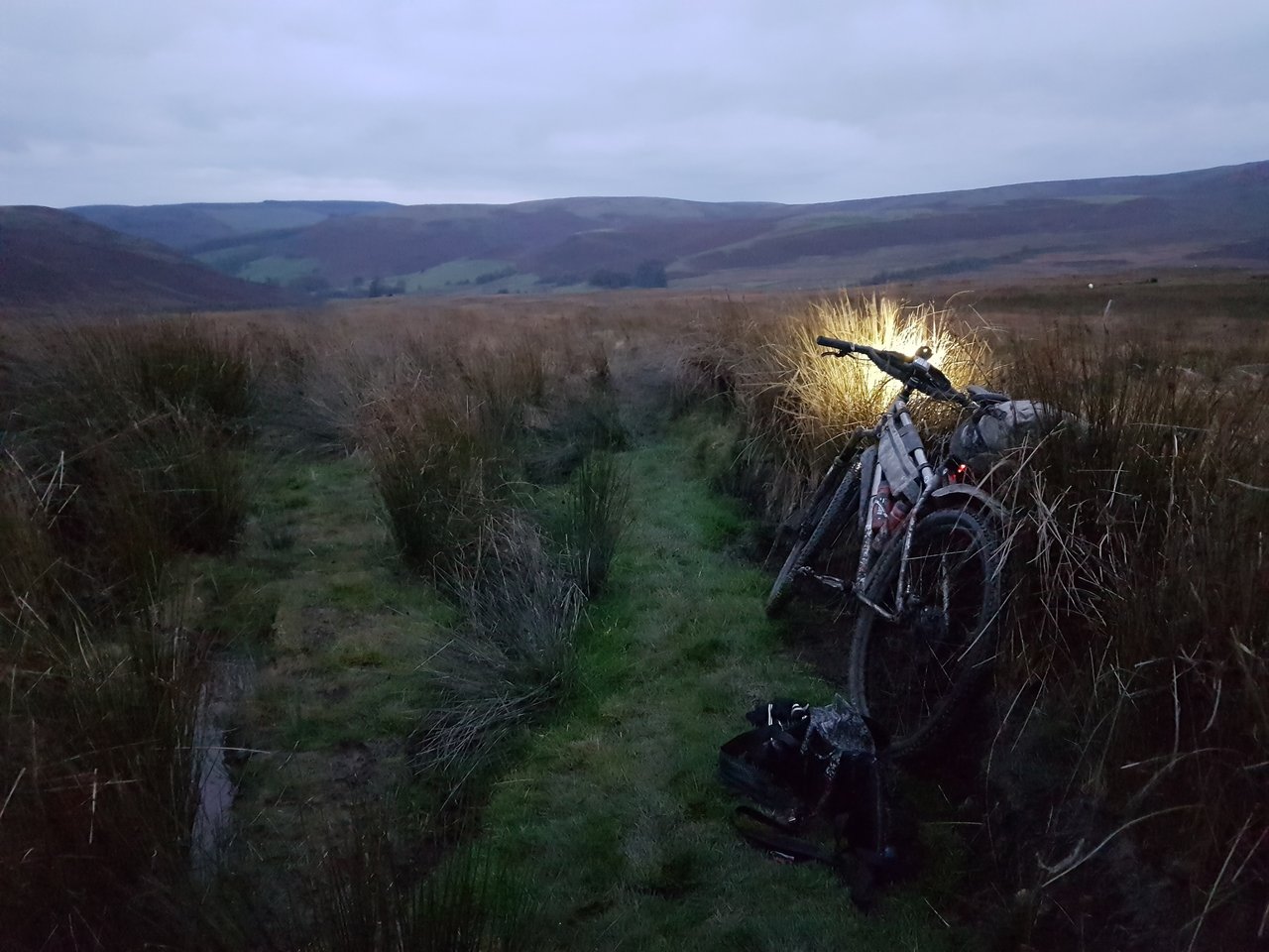Loaded bikepacking bike leaning in rushes beside a grassy Mid-Wales moorland track at dusk, lit by its front light.