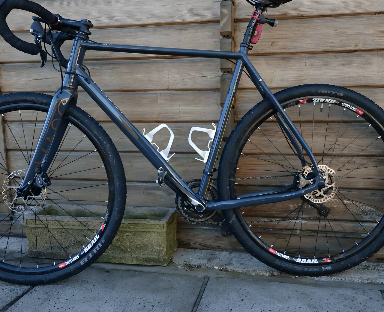 Dark grey Mason Bokeh gravel bike with WTB tyres and white bottle cages leans against a wooden shed wall.