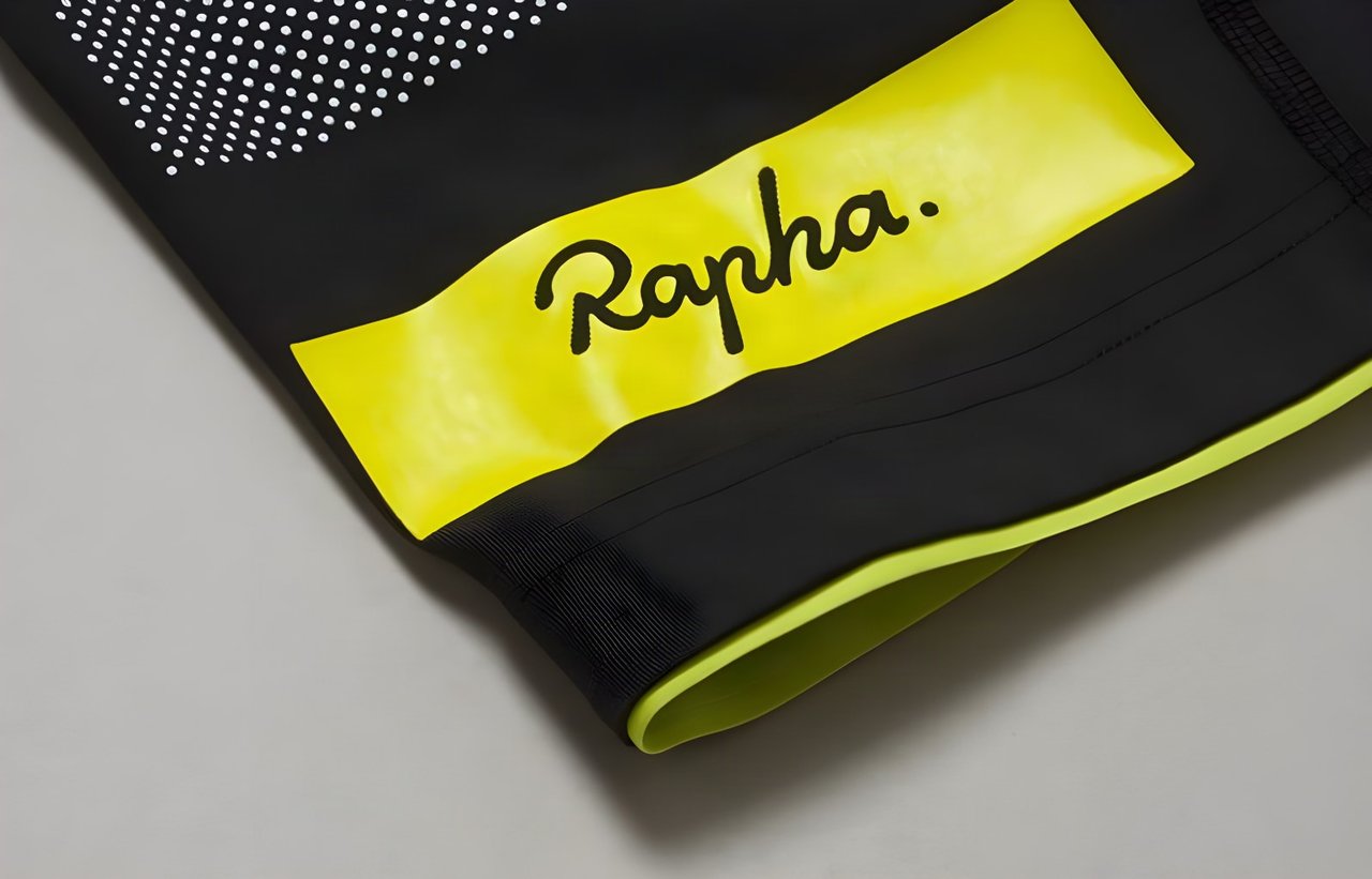 Close-up of a Rapha Brevet bib shorts leg hem with a yellow logo panel, white dots and yellow gripper edge on black fabric.