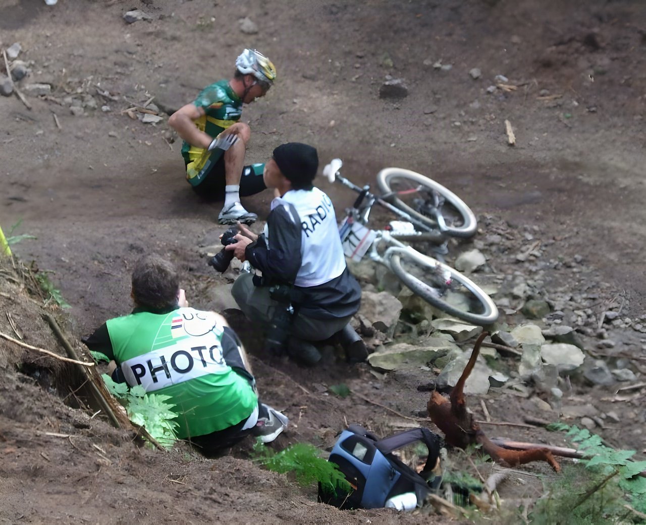 Mountain bike racer sits on the ground after a crash while two photographers crouch nearby and the bike lies on rocks.