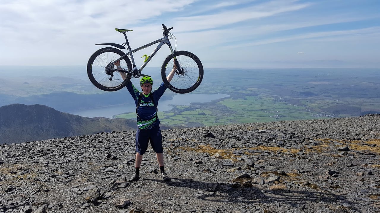 James Pearson holds his Whyte hardtail overhead on a stony Lake District summit, with a lake and fields far below.