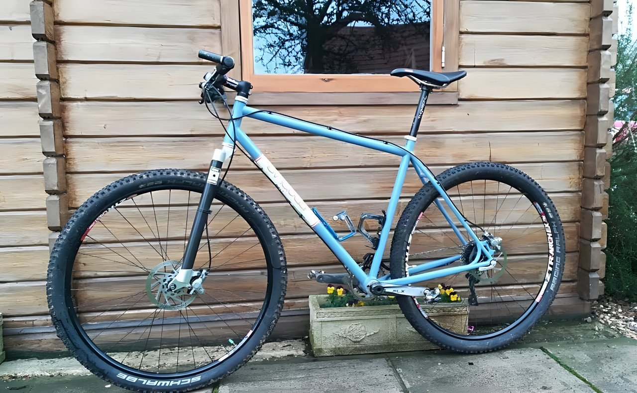 Singular Swift rigid steel 29er in pale blue leaning against a wooden cabin wall, fitted with On One carbon forks.