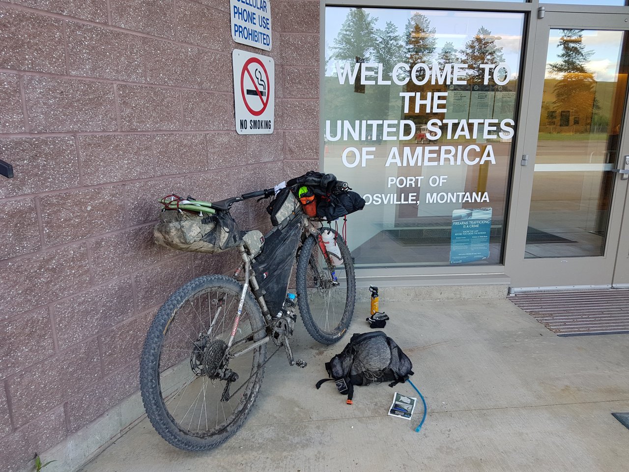 Mud-caked bikepacking bike outside the US port of entry at Roosville, Montana, with kit laid out on the ground.