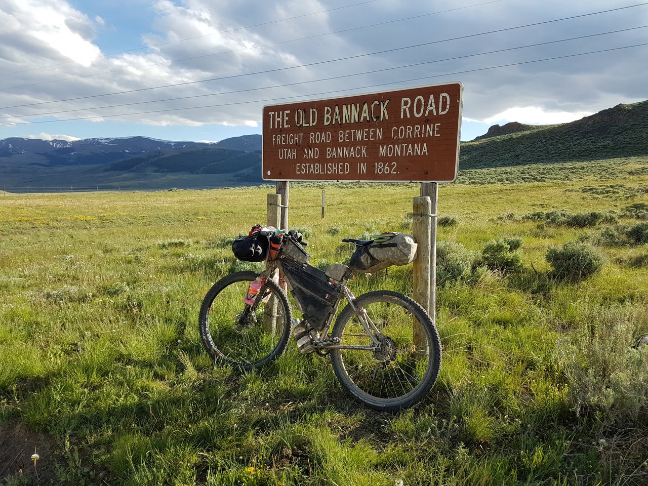 Loaded bikepacking bike leaning on The Old Bannack Road sign on the Tour Divide route, with mountains behind.