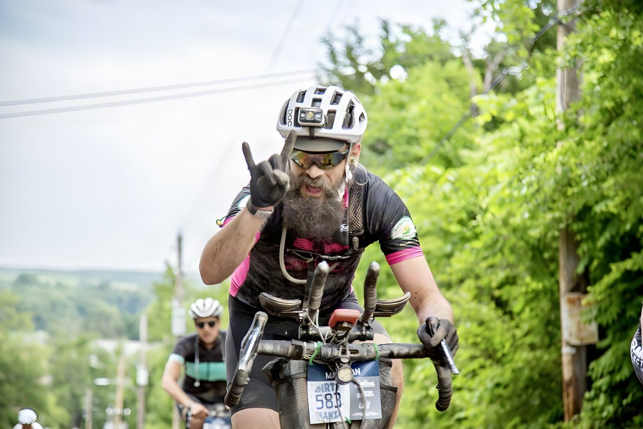 David Markman, a bearded Dirty Kanza 200 rider, climbs a hill on aero bars and flashes a hand sign at the camera.