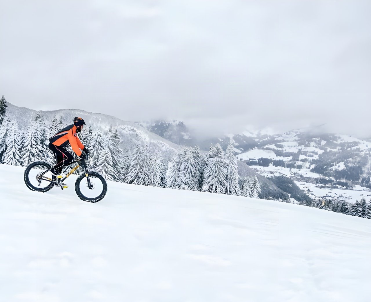 Fatbike rider in orange climbs a snowy slope above a valley on Stage 1 of the Snow Bike Festival in Switzerland.