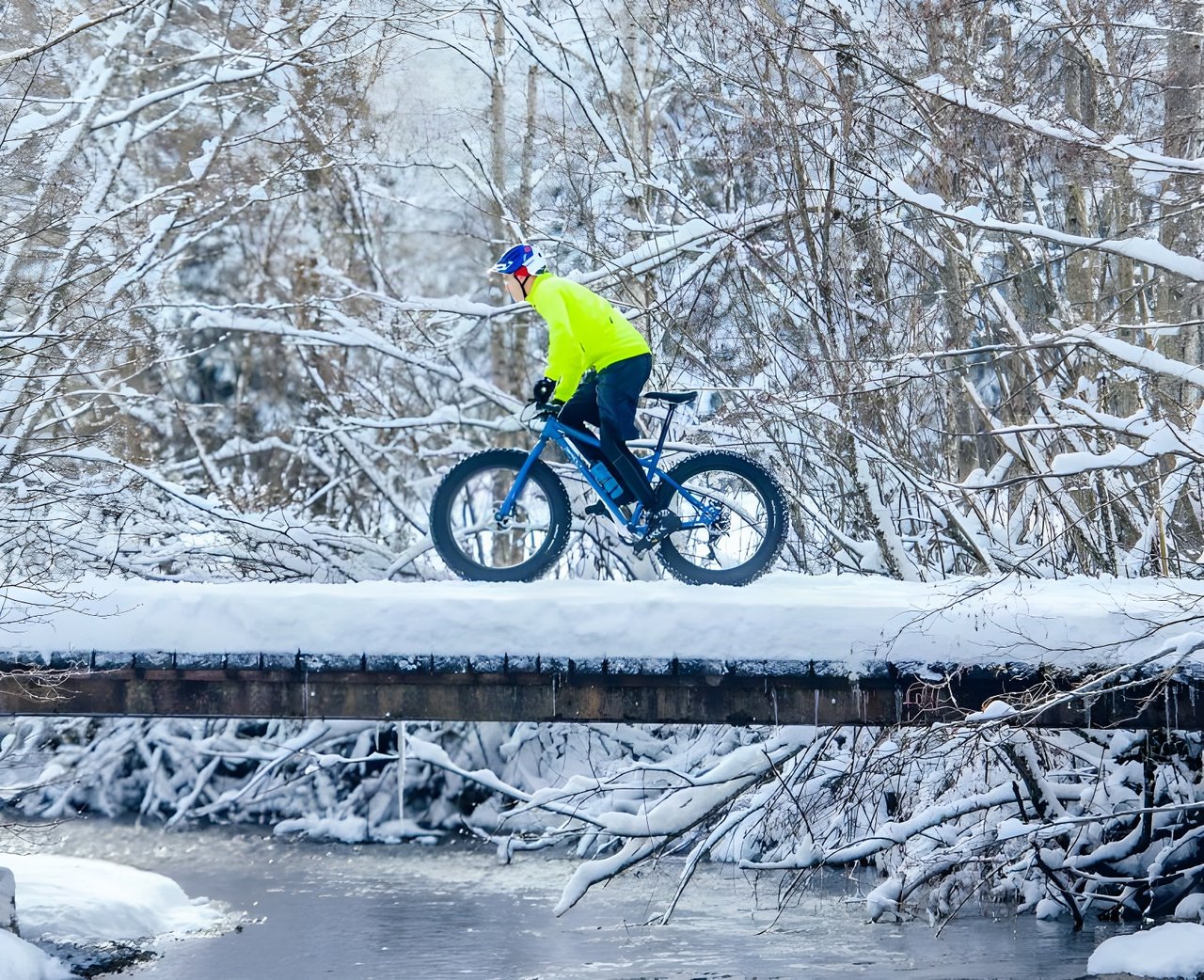 Rider in a yellow jacket crosses a snow-covered bridge on a blue fatbike during Stage 2 of the Snow Bike Festival.