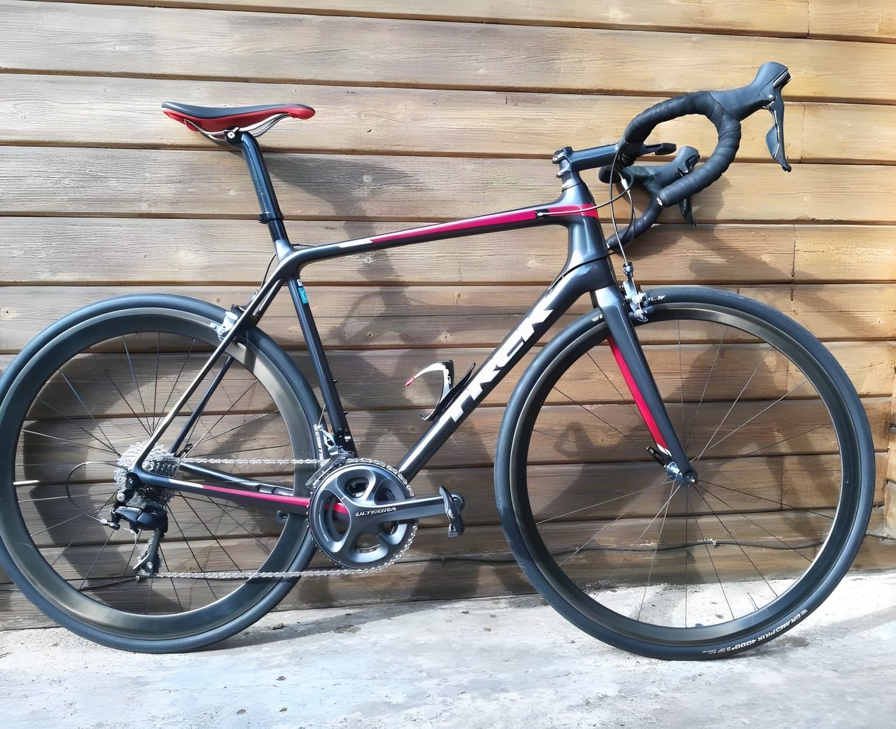 Trek Emonda SL road bike in charcoal and red with Shimano Ultegra and deep carbon wheels, against a timber wall.
