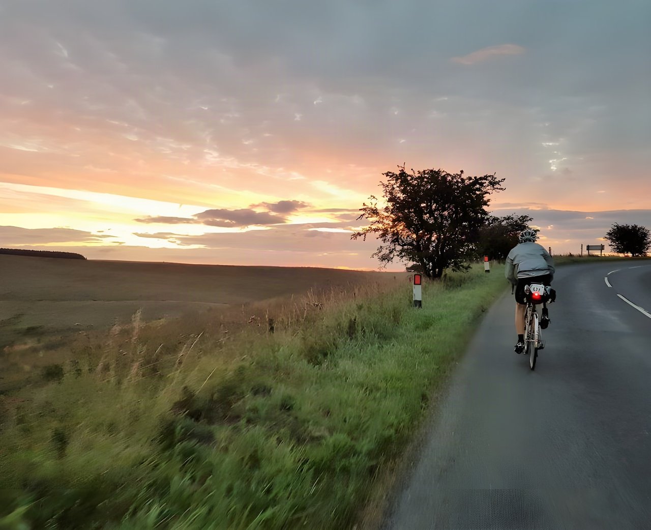 London-Edinburgh-London rider cycles a quiet country road at sunrise under a pink and orange sky.