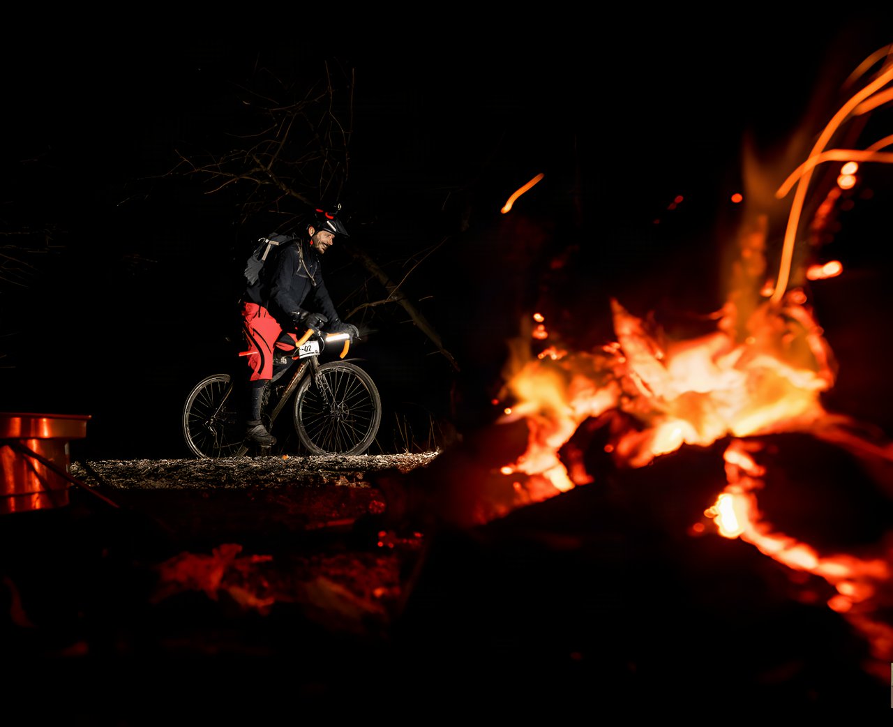 Rider with a helmet light stands astride a gravel bike beside a bonfire at night at Yorkshire True Grit Dark Skies.