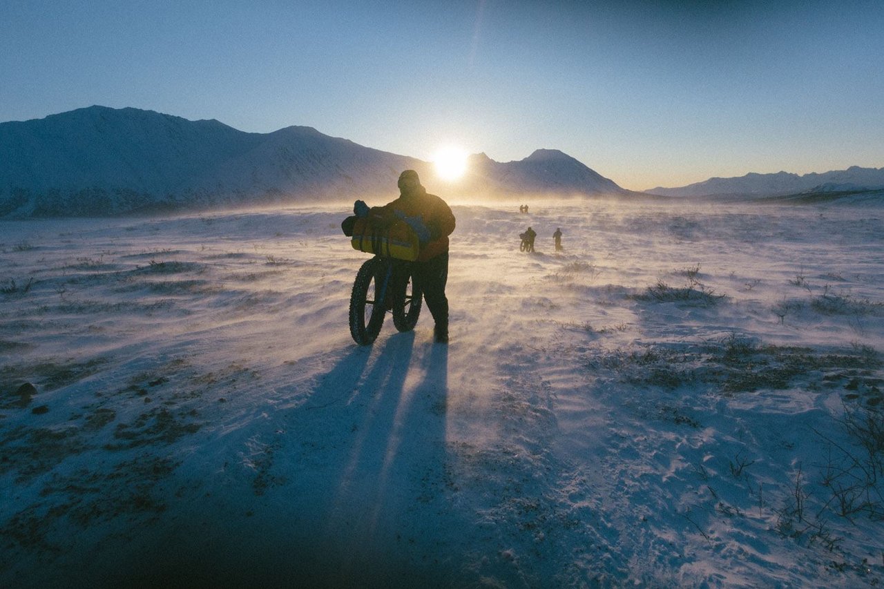 Fat bike rider pushes a loaded bike through wind-blown snow under a low sun on the Iditarod Trail in Alaska.