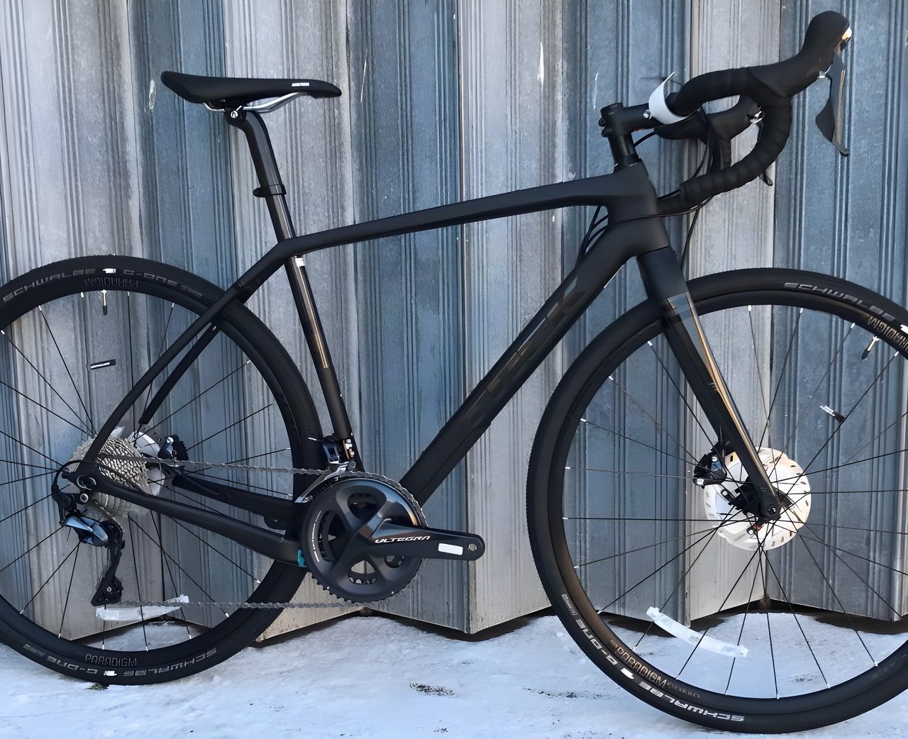 Matt black Trek Checkpoint gravel bike with Shimano Ultegra cranks and Schwalbe tyres, leaning on a metal wall.