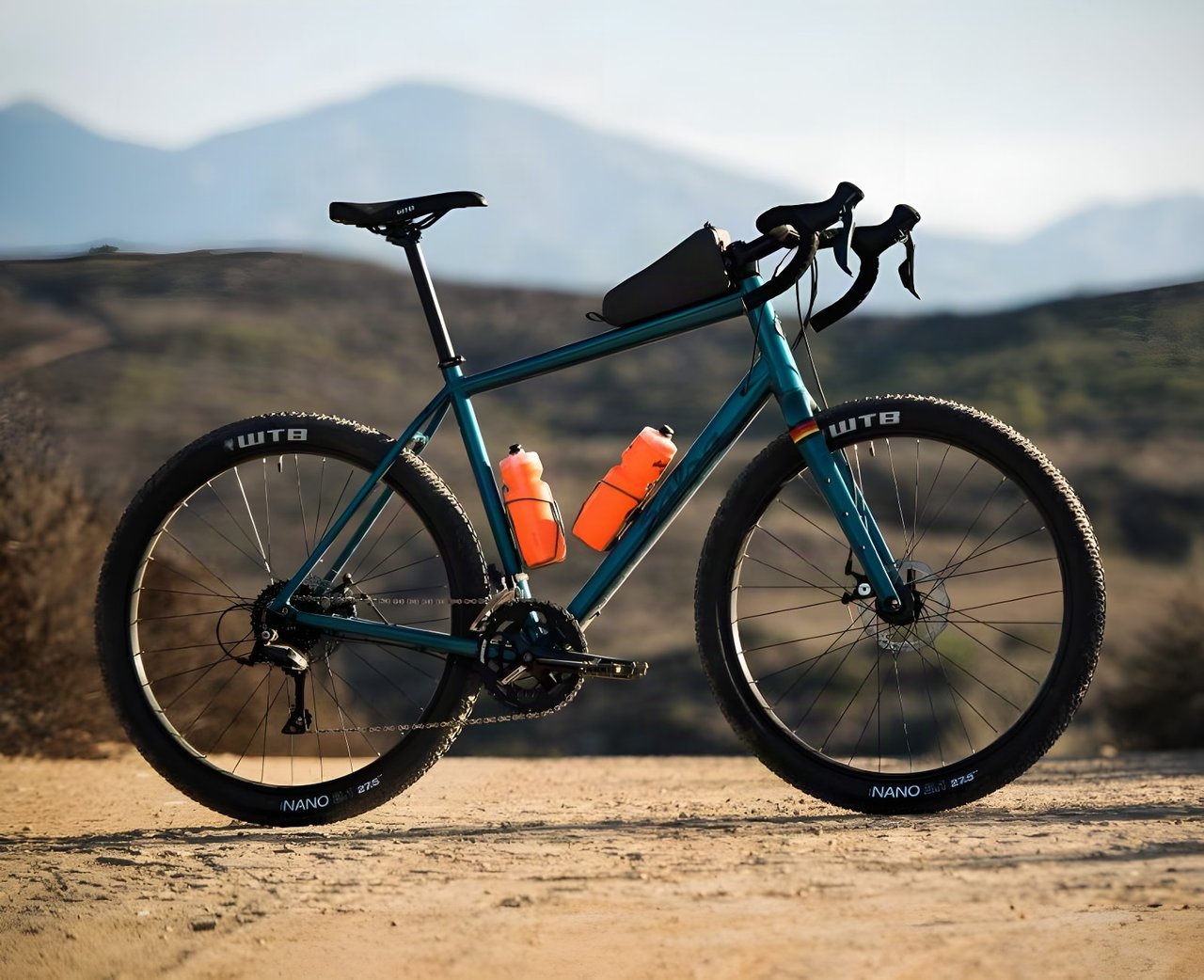 Salsa Journeyman gravel bike in teal with orange bottles, WTB Nano 27.5 tyres and a top tube bag, on dusty ground.