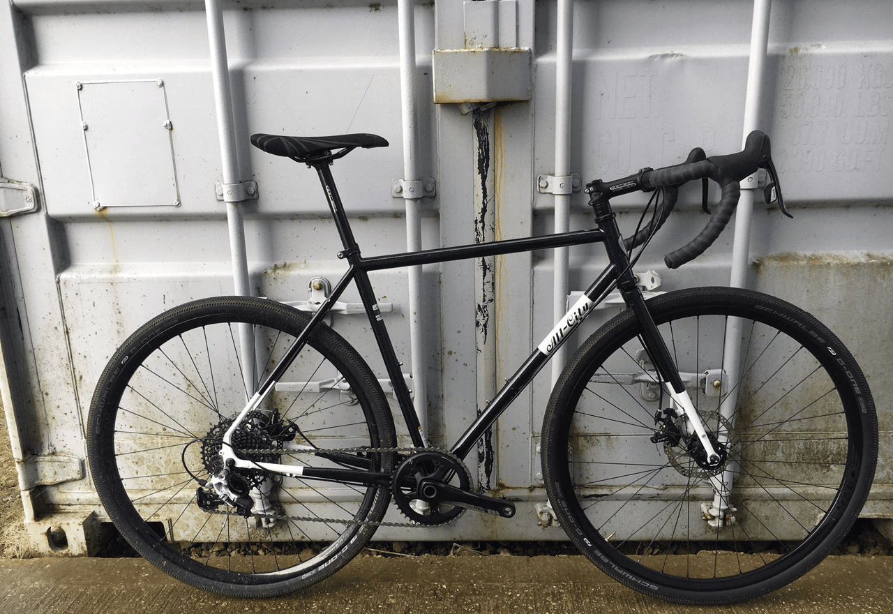 Black All-City Macho Man steel cyclocross bike with 1x drivetrain and disc brakes leaning on a shipping container.
