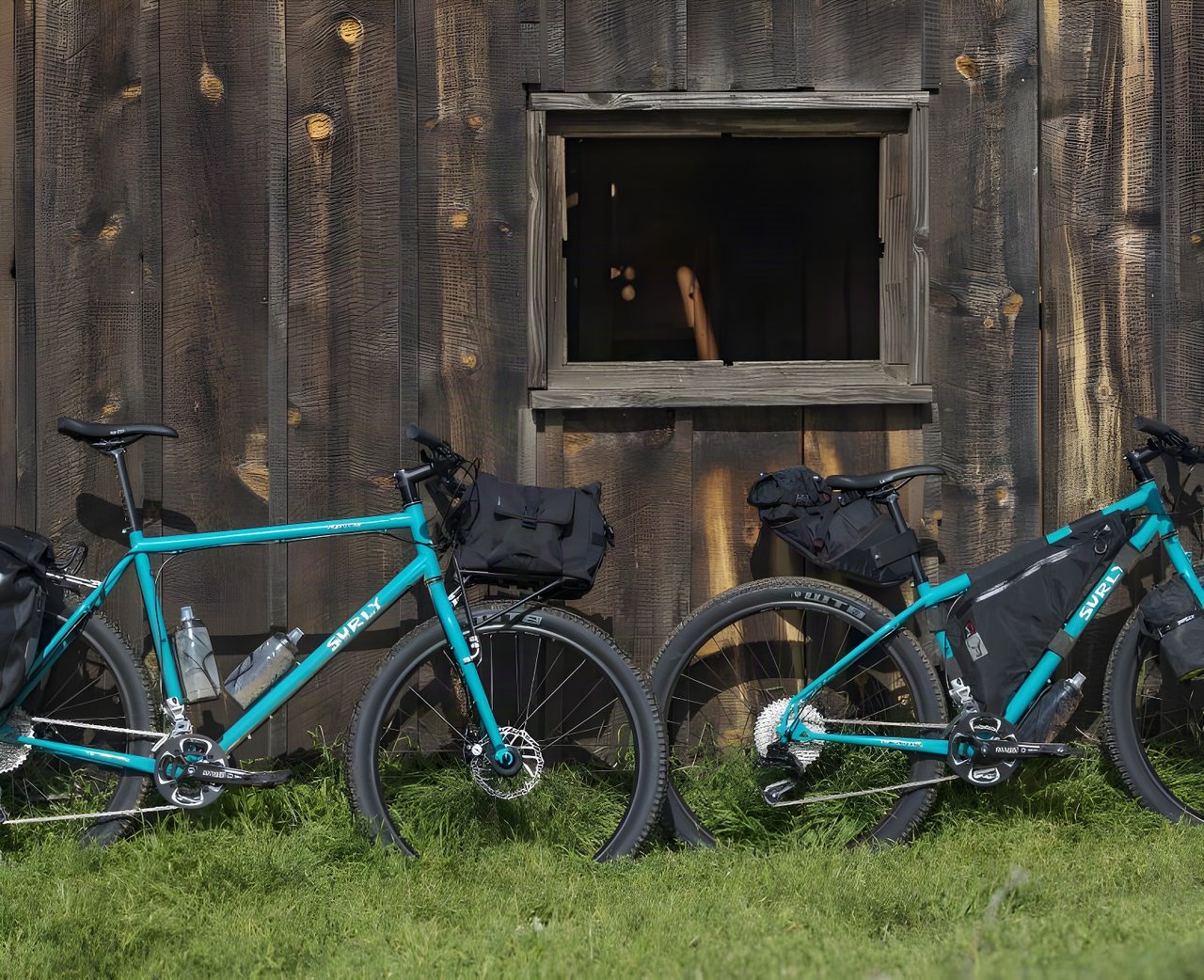 Two teal Surly Bridge Club bikes loaded with bikepacking bags, leaning against a weathered wooden barn.
