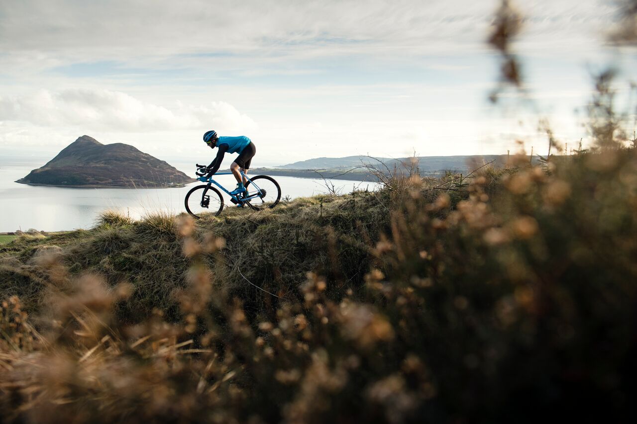 Grinduro Scotland rider on a blue gravel bike crests a heather ridge above the sea on the Isle of Arran.