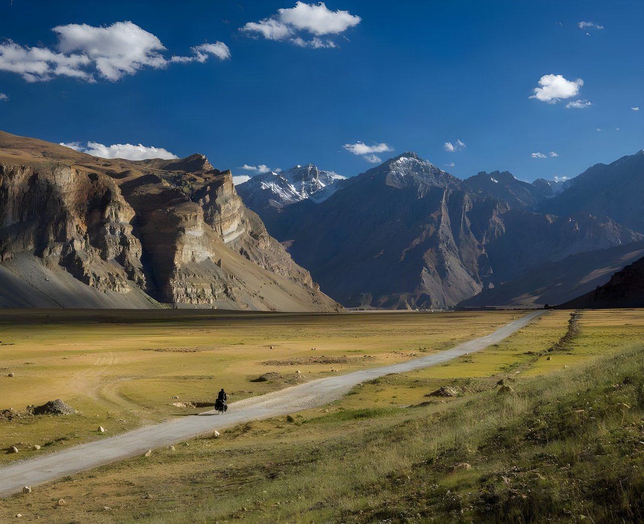 Cyclist rides a gravel road across a high valley beneath mountains, the cover image of Escape By Bike by Joshua Cunningham.