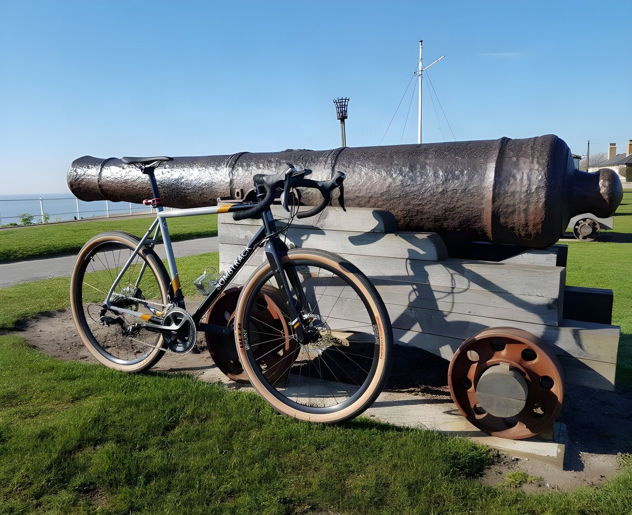 Bombtrack Audax steel road-plus bike with tan-wall tyres leaning on a cannon on a clifftop green.