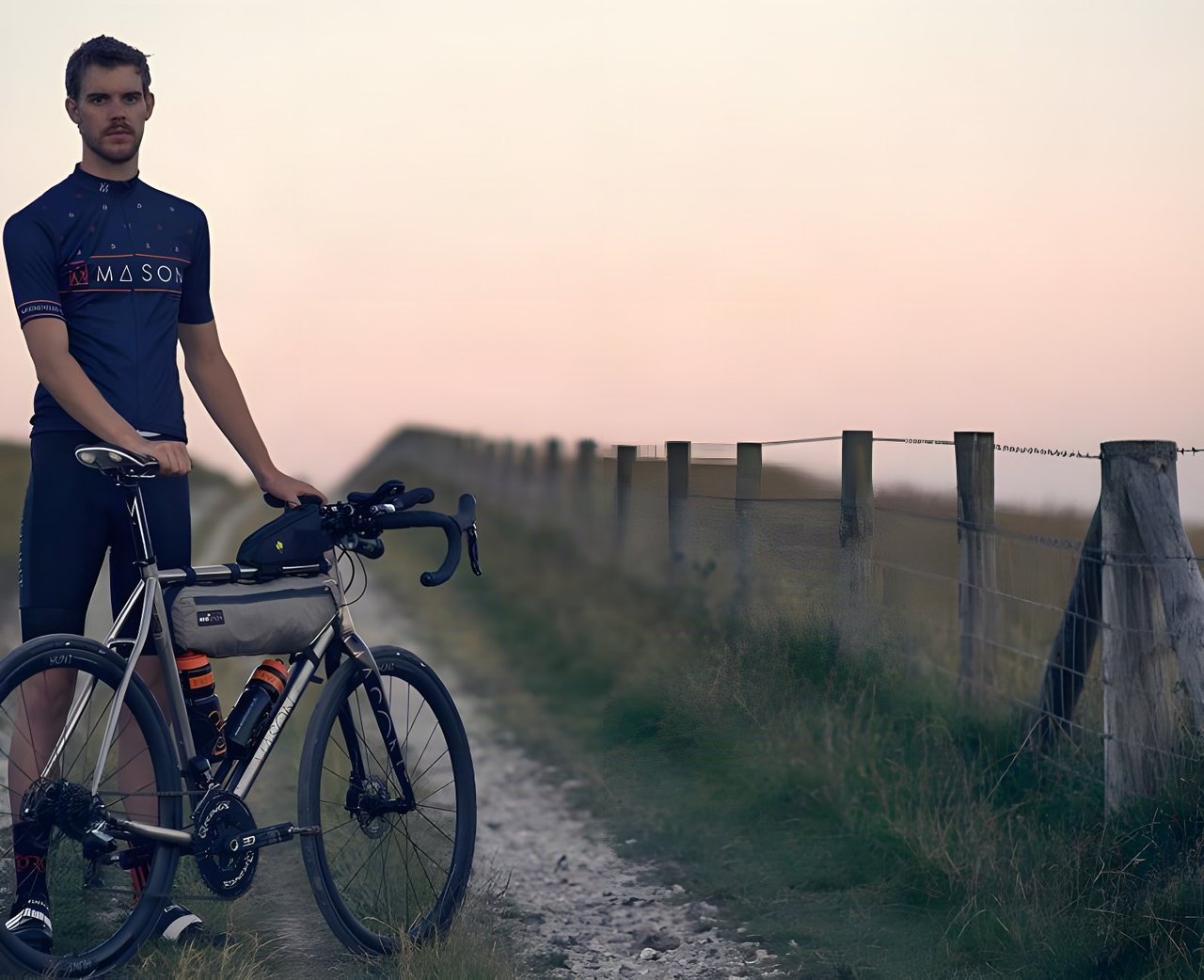 Josh Ibbett, 2015 Transcontinental Race winner, stands in a Mason jersey beside his Mason bike on a chalk track at dusk.