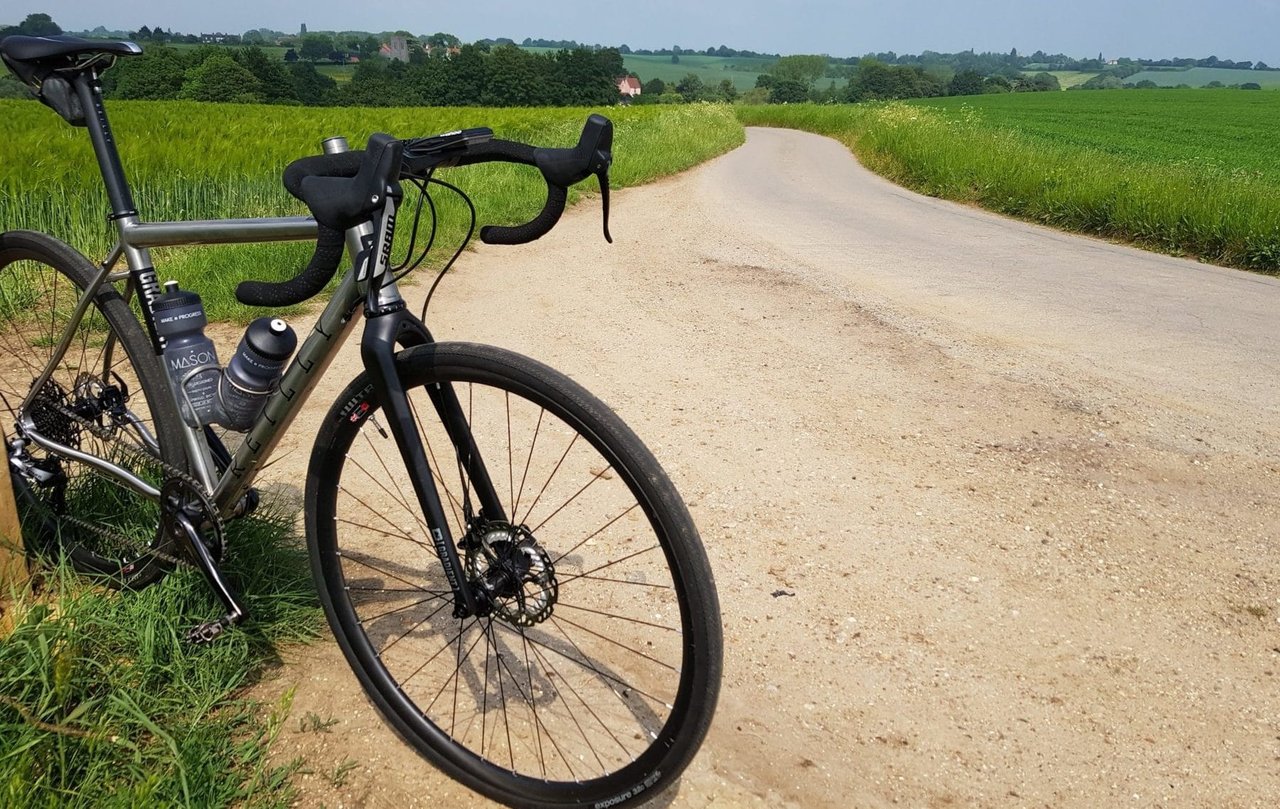 Reilly Gradient titanium adventure bike propped at the edge of a gravel lane between green fields.