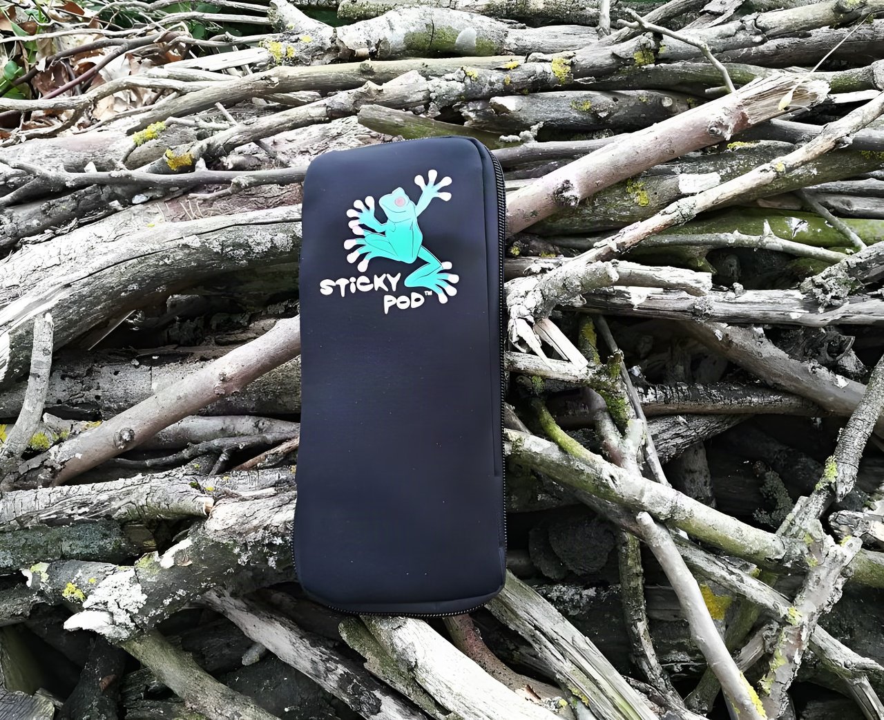 Black neoprene Large Sticky Pod with its green frog logo lying on a pile of lichen-covered sticks.