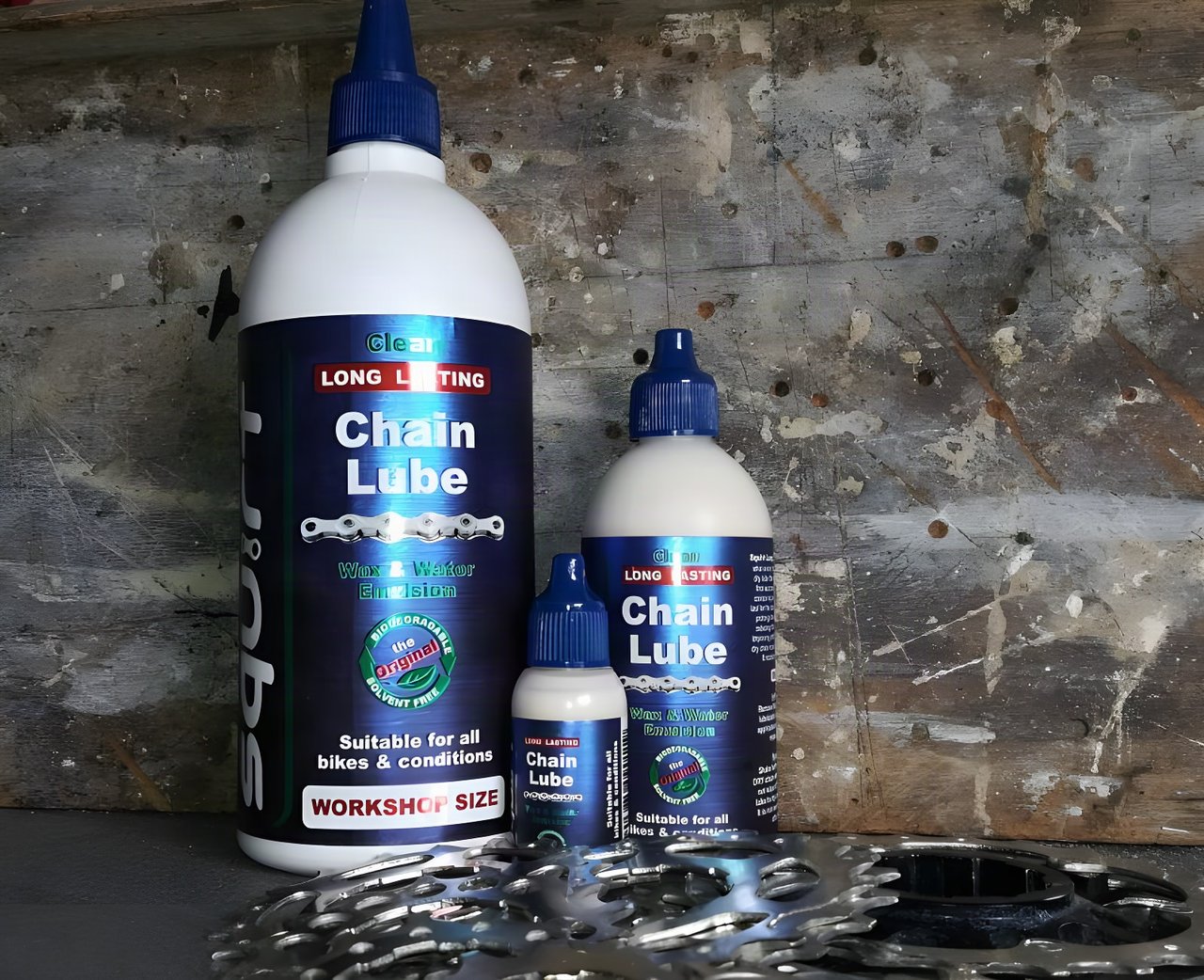 Three sizes of Squirt Long Lasting Chain Lube, a wax and water emulsion, lined up with a cassette against weathered wood.