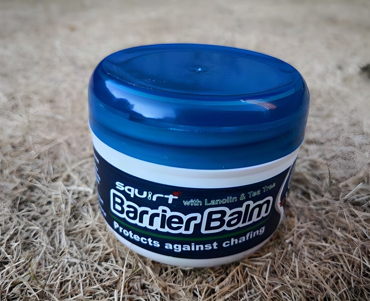 Blue tub of Squirt Barrier Balm chamois balm, labelled with lanolin and tea tree, sitting on dry grass.