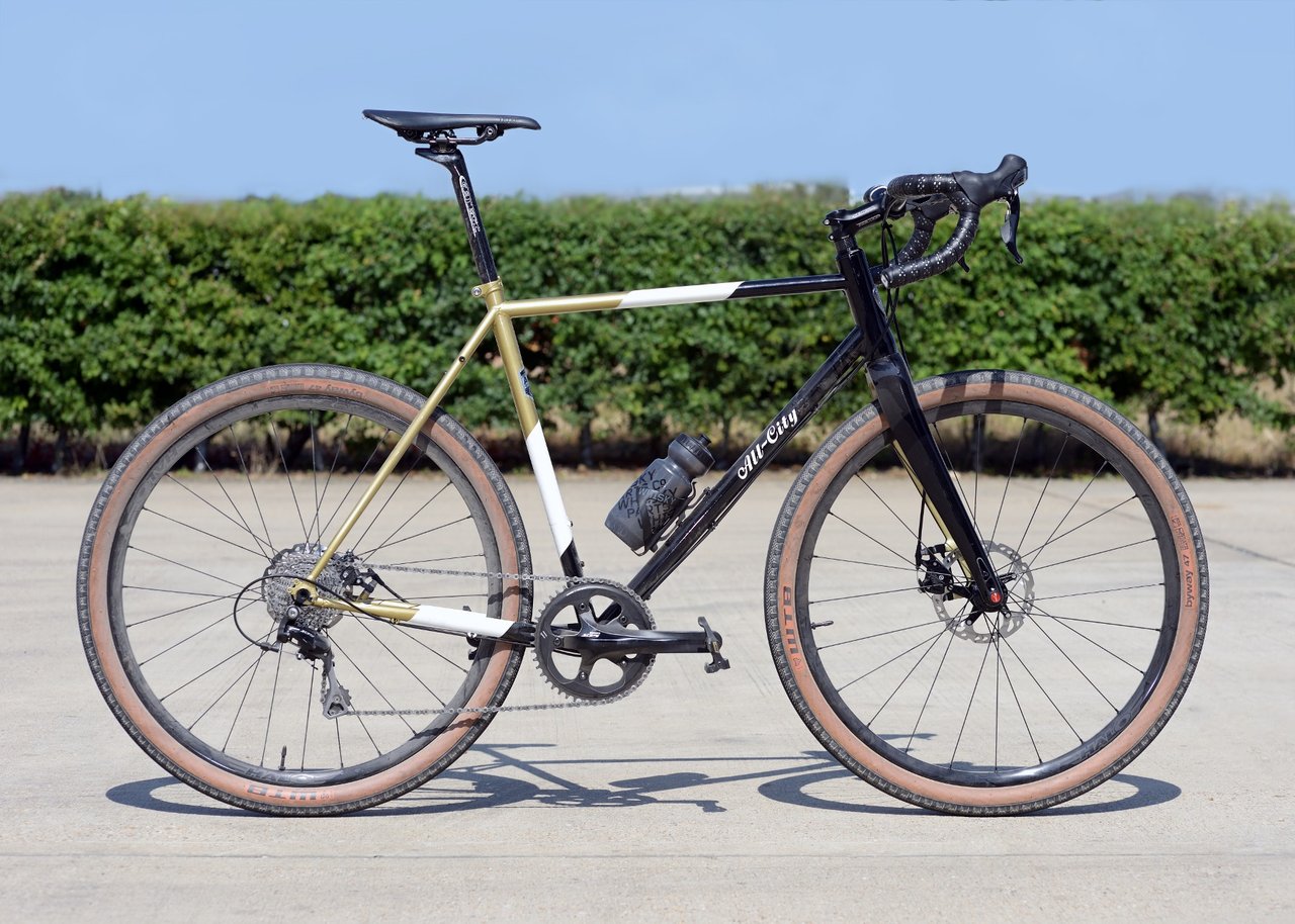 Jordan Lunn's gold and black All-City Cosmic Stallion gravel bike with tan-wall WTB tyres, side on in the sun.