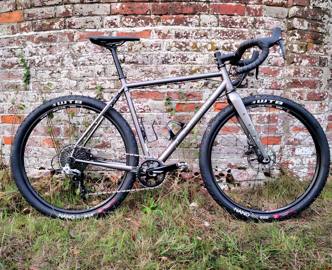 Bombtrack Hook EXT gravel bike in profile, matte grey steel frame with WTB Nano tyres, against a brick wall.