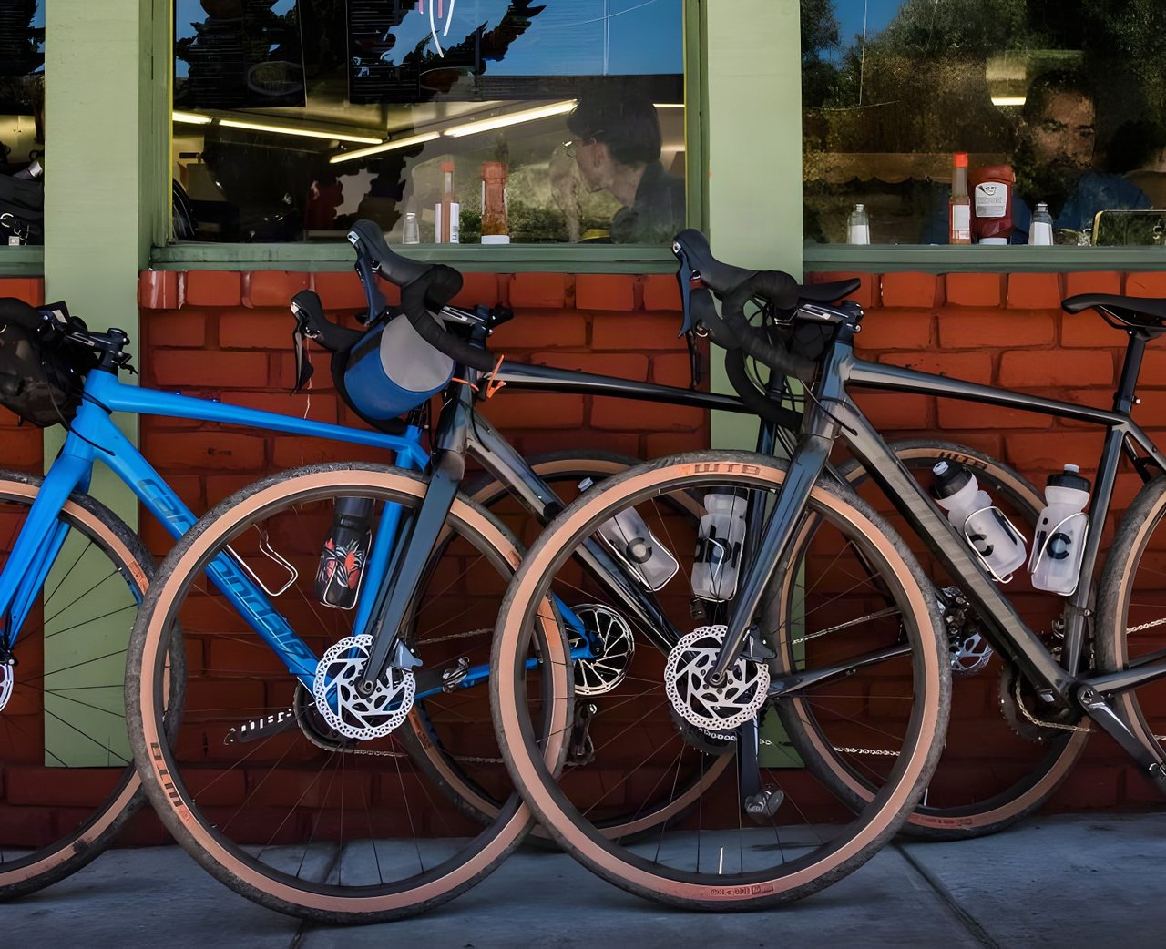 Three Cannondale Topstone gravel bikes, one blue and two grey, leaning on a brick wall below a café window.