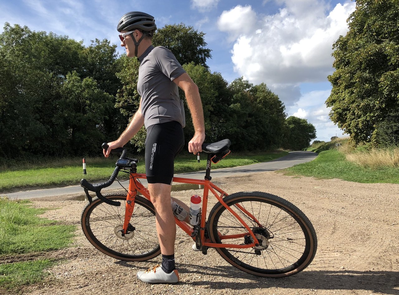 Rider in Endura FS260-Pro bibshorts stands beside an orange gravel bike on a gravel track by a country road.