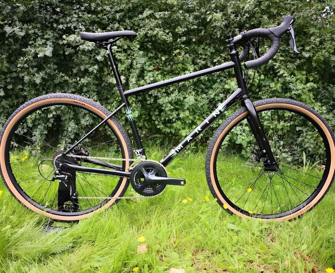 Black Marin Four Corners steel gravel bike with tan-wall tyres and drop bars, side view on grass in front of a hedge.
