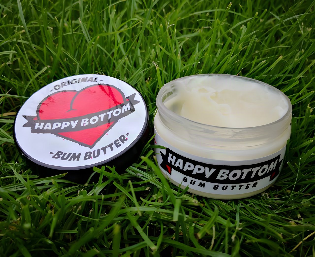 An open tub of Happy Bottom Bum Butter beside its lid on grass