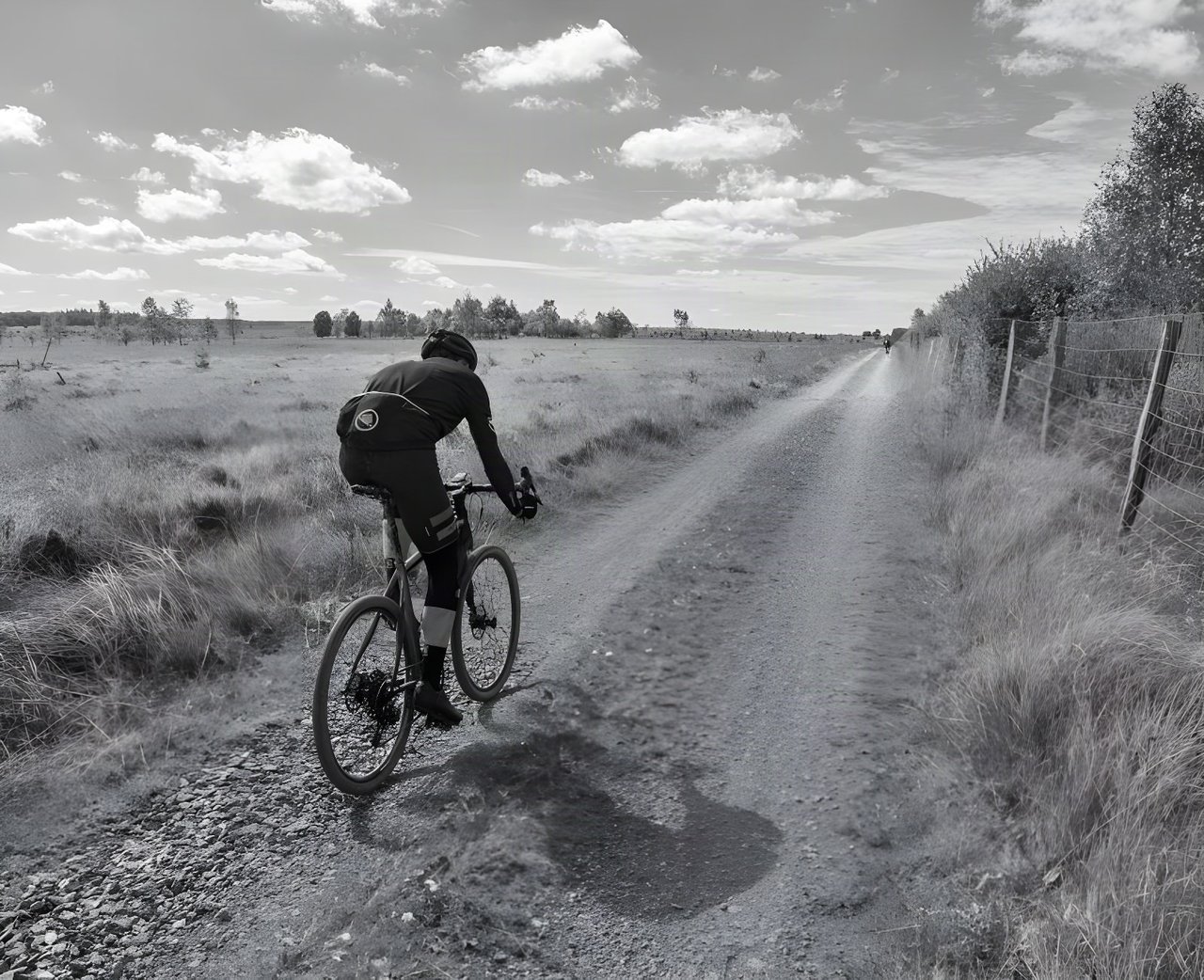 Rider on a gravel bike on a fenced heathland track at the Dirty Boar in Belgium, in black and white.
