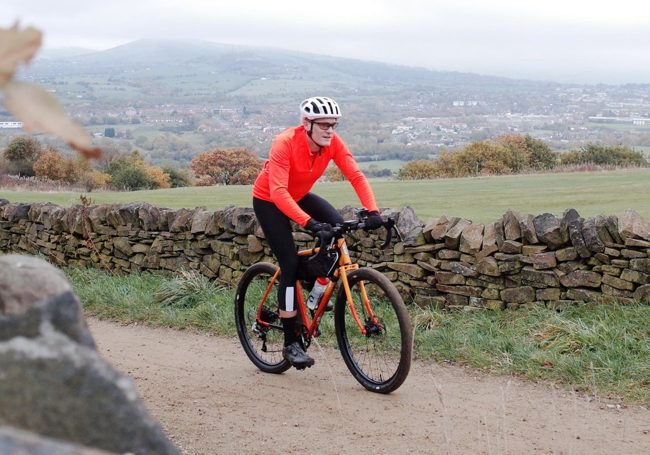 Rider in a bright orange POC AVIP Ceramic Thermal Jersey on a gravel bike beside a dry-stone wall.