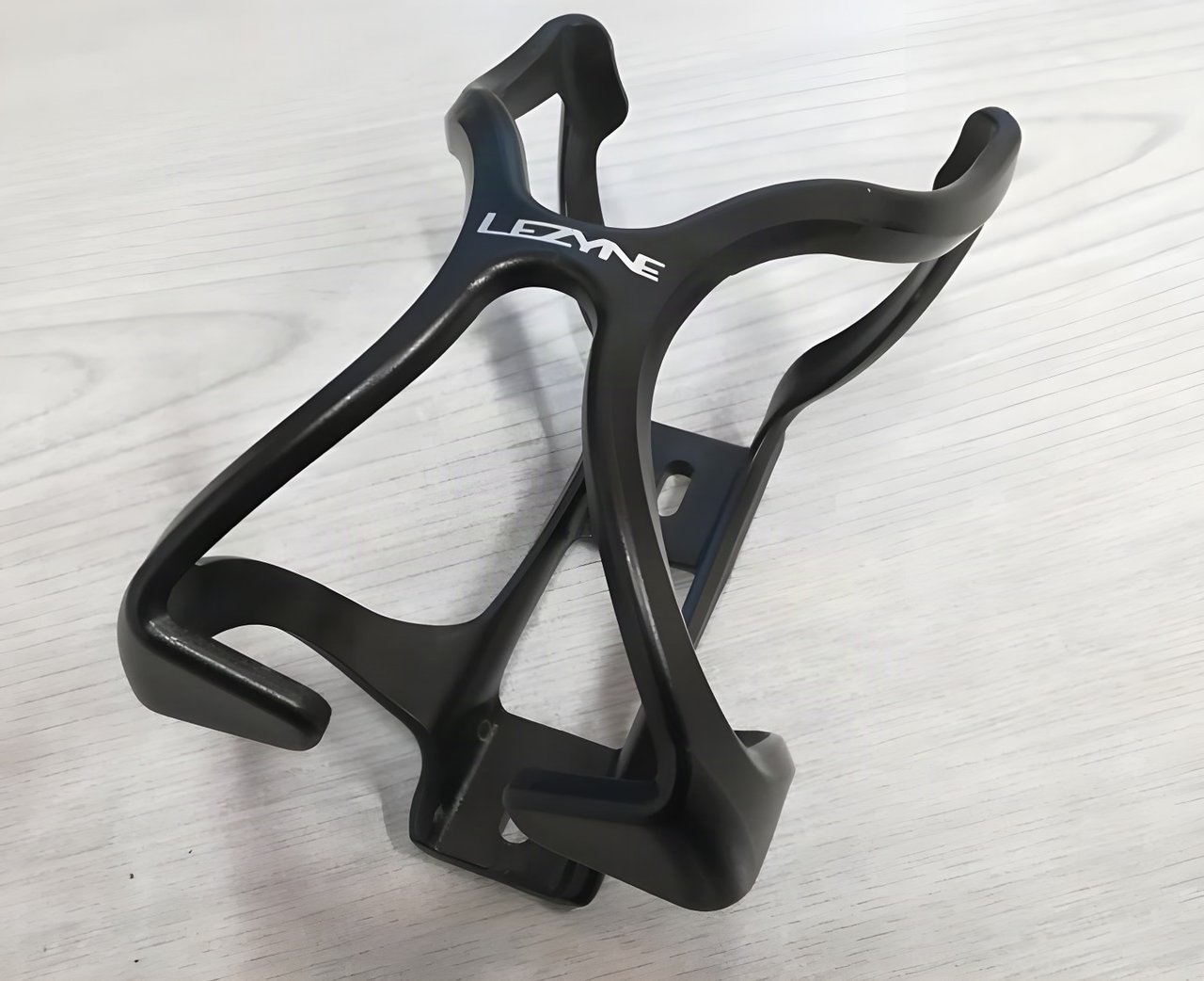 Lezyne Flow Cage in black, a plastic bottle cage with the brand logo, resting on a pale wooden surface.