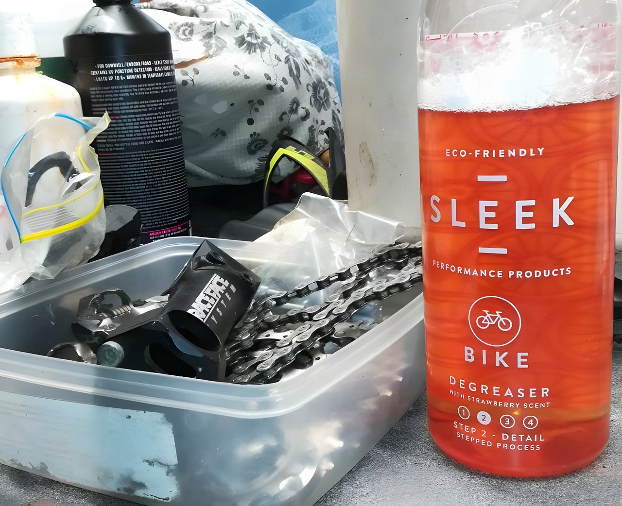 Bottle of SLEEK Bike Degreaser with strawberry scent beside a tub of bike chains and parts.