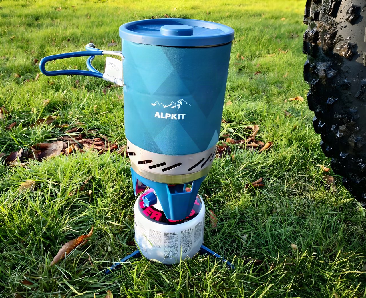 Alpkit BruKit stove with its blue 1-litre pot on a gas canister and tripod on grass, a muddy bike tyre alongside.