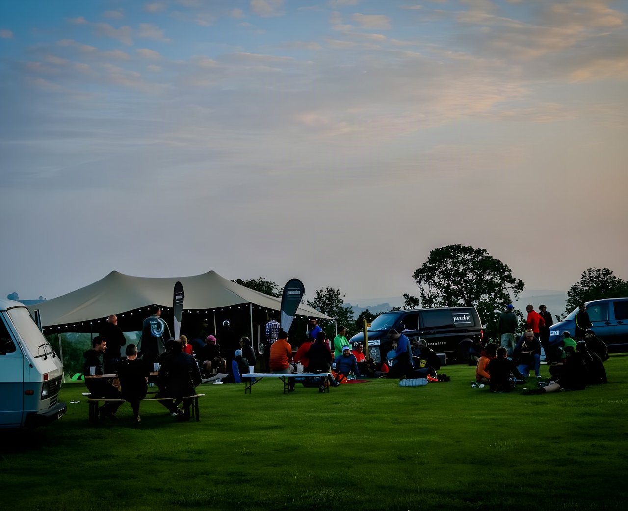 Riders sit on the grass at The Distance gravel event base camp beside a marquee and vans at dusk.