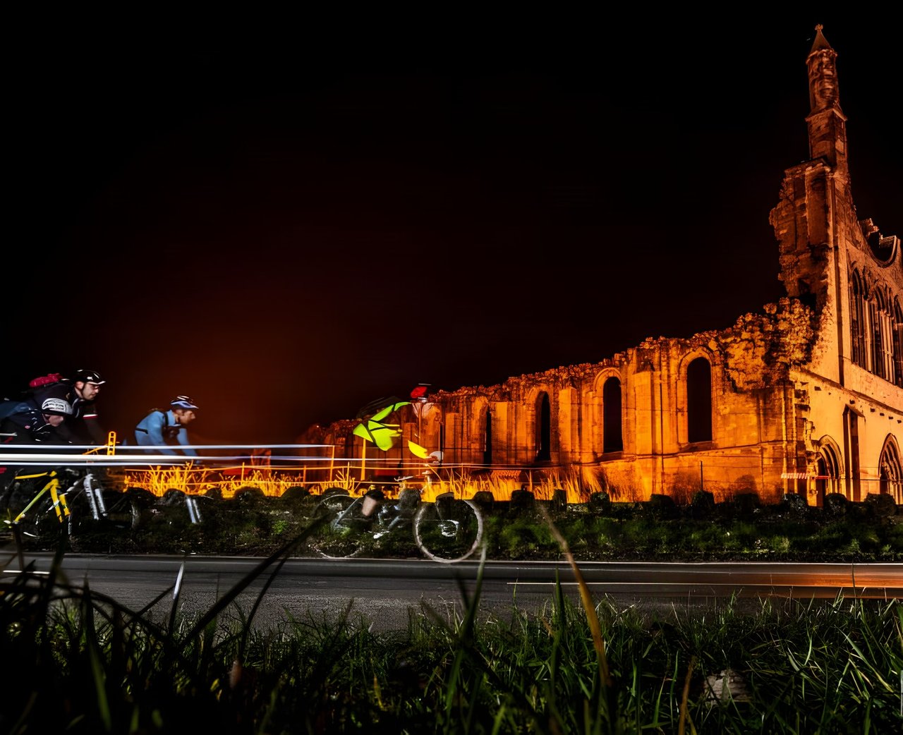 Riders with lights streak past floodlit stone ruins at night during Yorkshire True Grit Dark Skies.