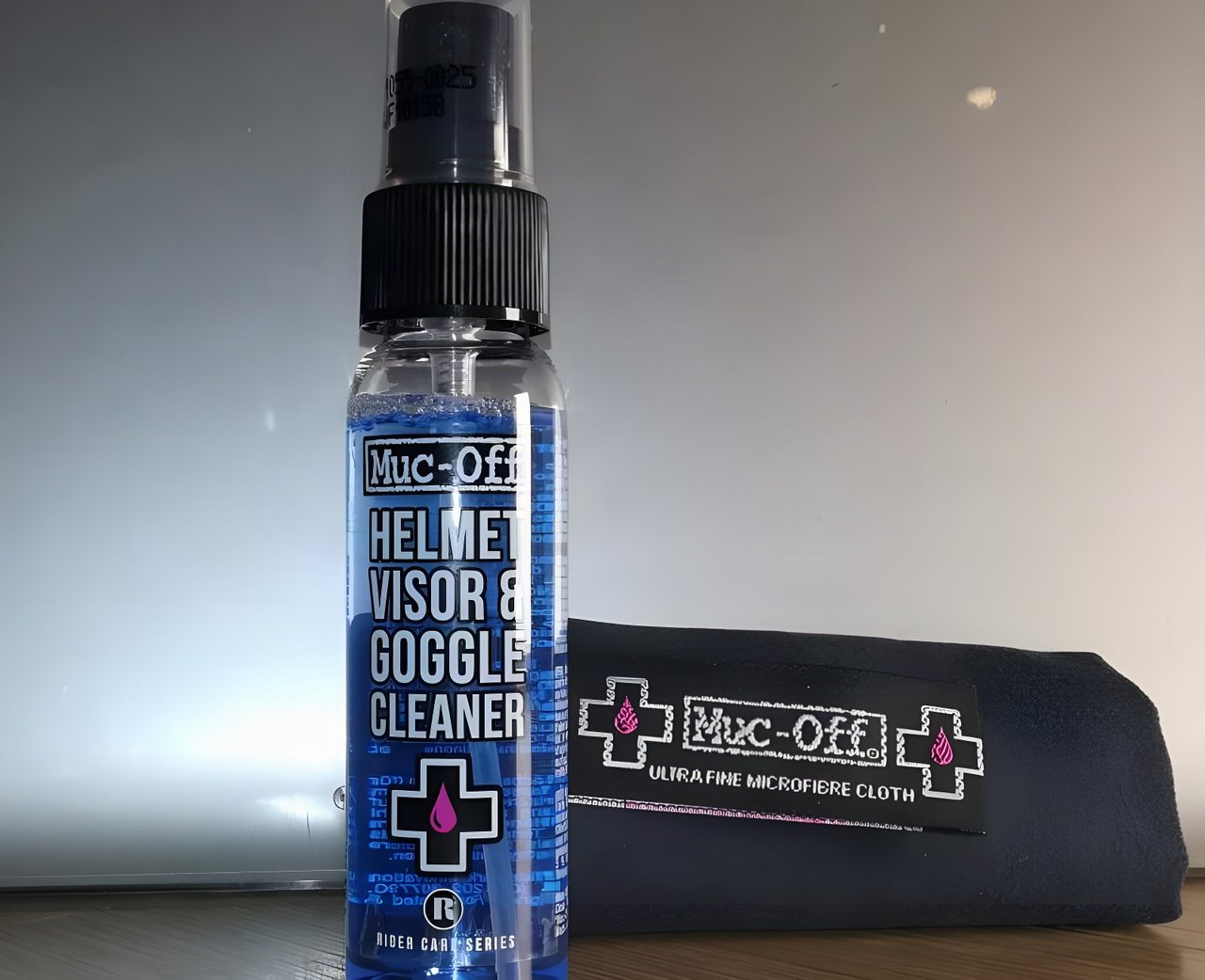 Muc-Off Helmet, Visor and Goggle Cleaner spray bottle beside its ultra fine microfibre cloth.