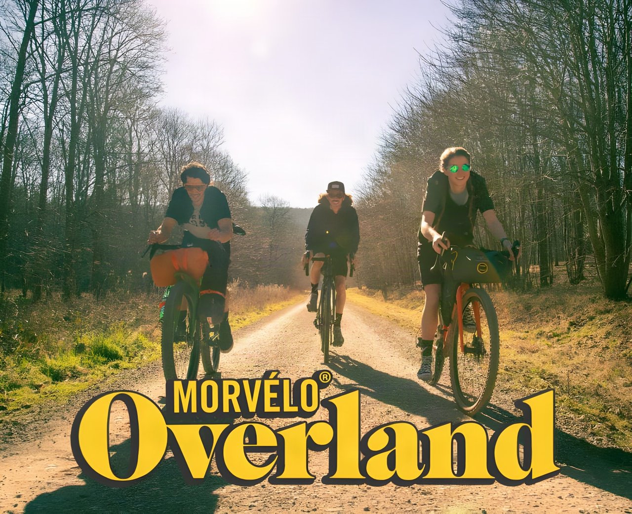 Three riders on a sunny gravel track with loaded bikes under the Morvélo Overland logo.