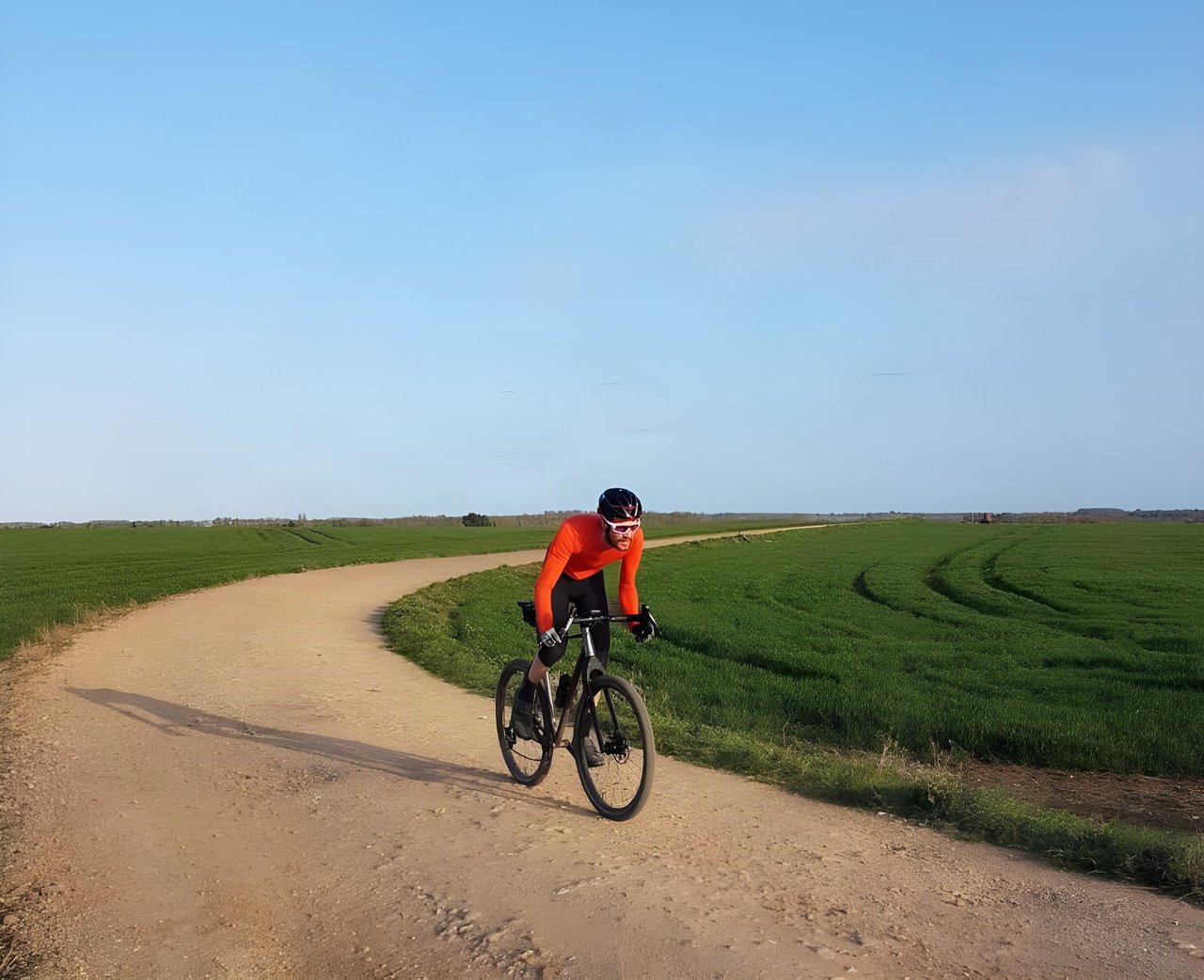 Josh Ibbett rides his Mason ISO along a curving gravel track through green fields under a clear blue sky.