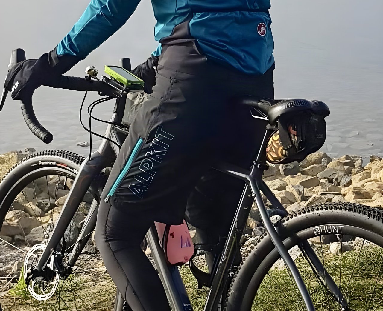 Rider in black Alpkit Floe women's MTB shorts with teal zip on a gravel bike beside water.