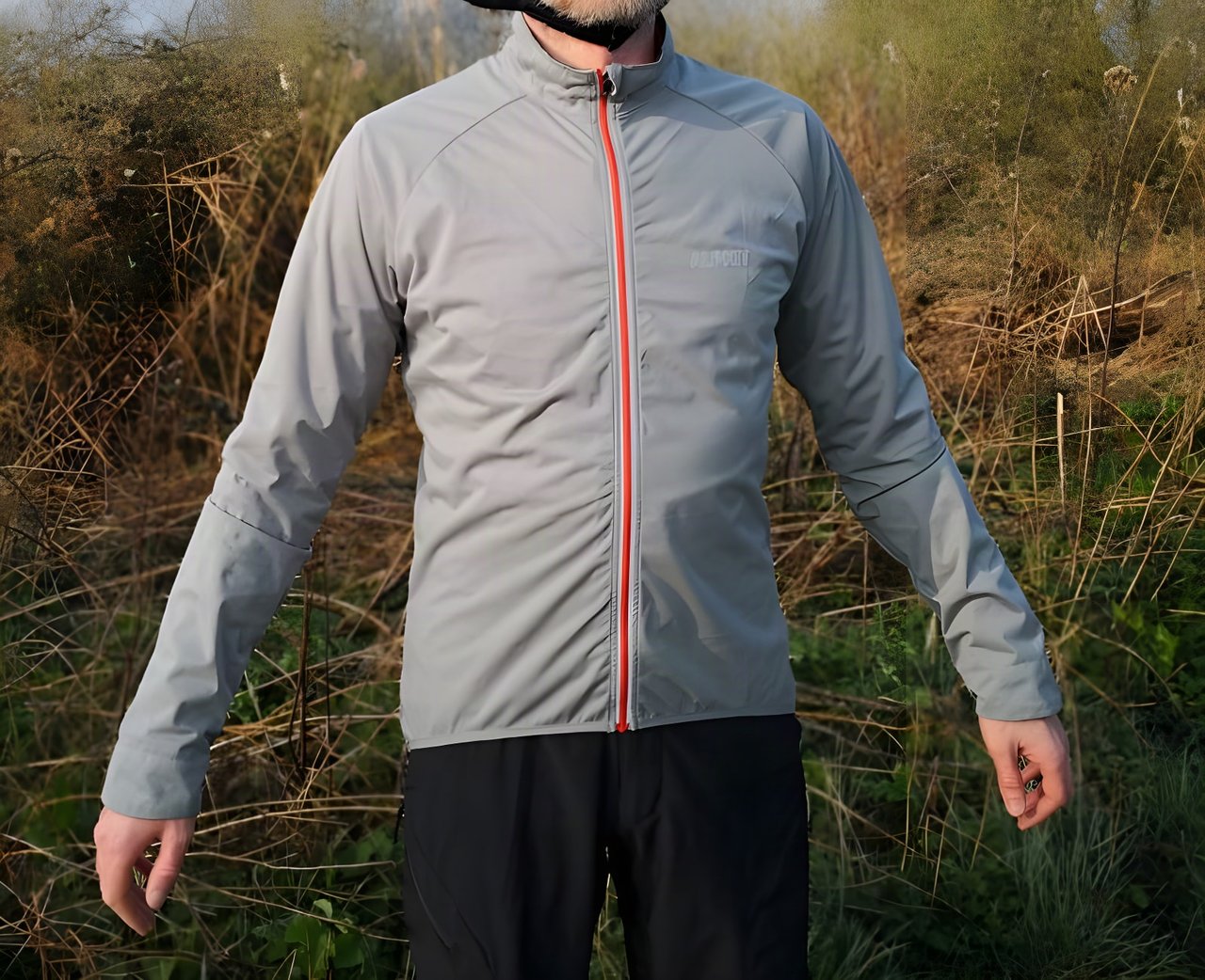 Alpkit Squalline jacket in steel grey with a red front zip, worn by a rider standing among dry winter grasses.