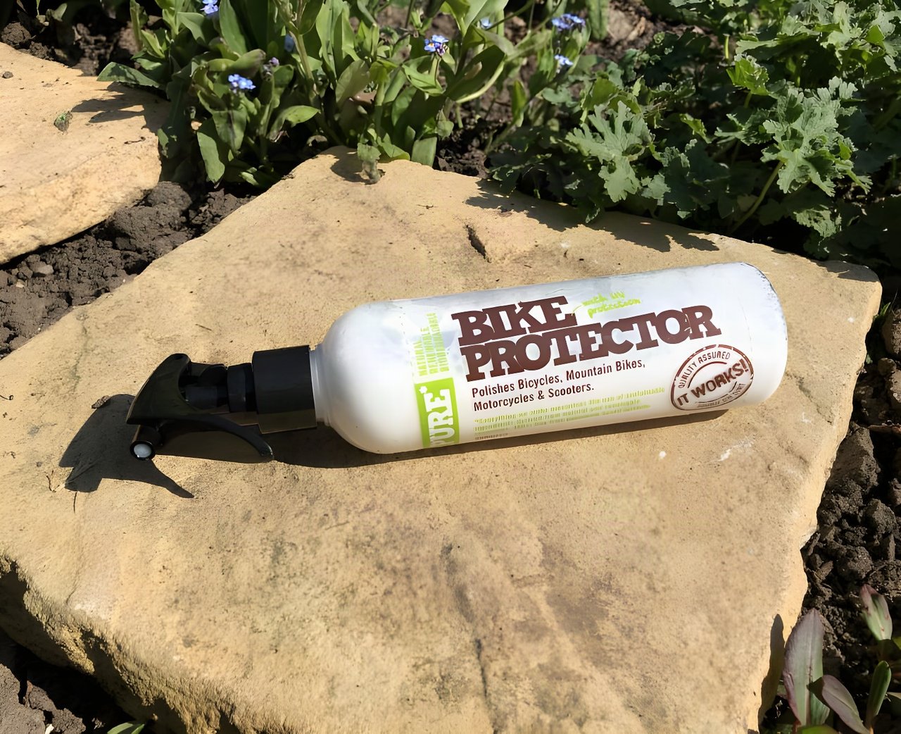 Weldtite Pure Bike Protector spray bottle lying on a flat stone beside garden plants.
