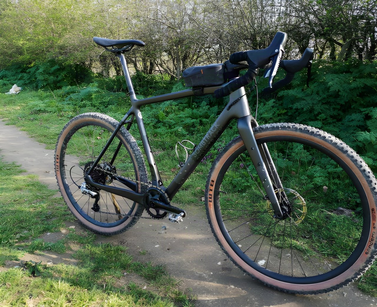 Bombtrack Hook EXT-C carbon gravel bike with a top-tube bag and tan-wall WTB Sendero tyres on a dirt track beside a hedge.