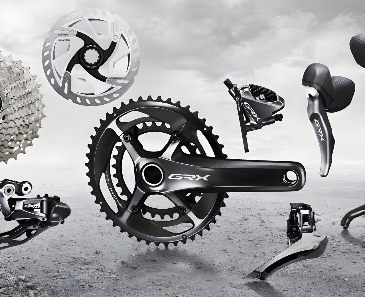 Shimano GRX gravel groupset components, including crankset, derailleurs, brake caliper, rotor and shifter, on grey gravel.