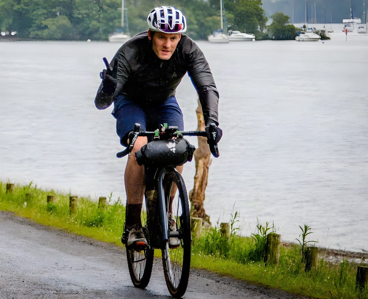 Rider in the black 7Mesh Oro jacket on a gravel bike with a bar bag, riding a wet lakeside road in the rain.