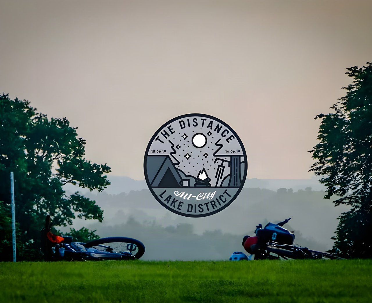 The Distance 2019 Lake District badge over two bikes lying on grass at dusk above a misty valley.